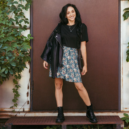 Woman in a floral skirt and black top standing in front of a brown door with plants around.