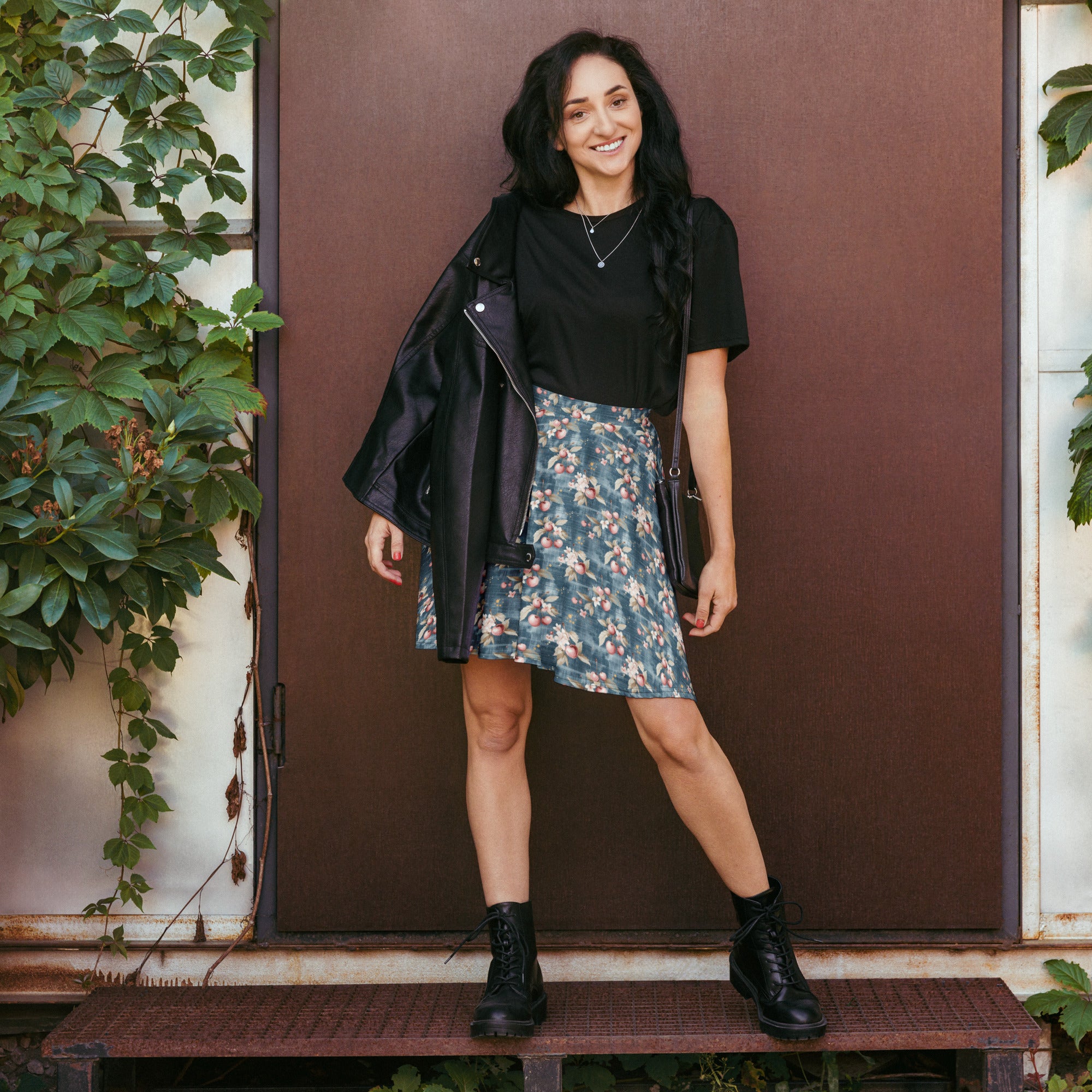 Woman in a floral skirt and black top standing in front of a brown door with plants around.
