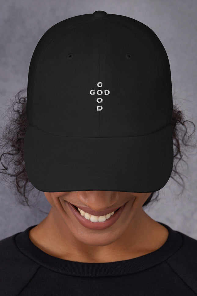 Person wearing a black cap with 'He is Good' embroidered on it against a gray background